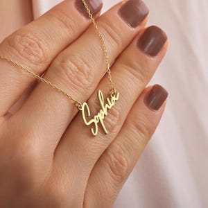 Pode incluir: Um colar dourado com o nome "Sophia" em fonte cursiva. O colar é segurado por uma pessoa com esmalte de unhas marrom. O fundo é uma camisa branca.