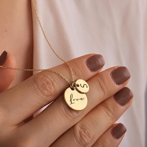 Może przedstawiać: Złoty naszyjnik z dwoma okrągłymi wisiorkami. Na większym wisiorku napis "Love", a na mniejszym litera "S". Naszyjnik ma delikatny łańcuszek.
