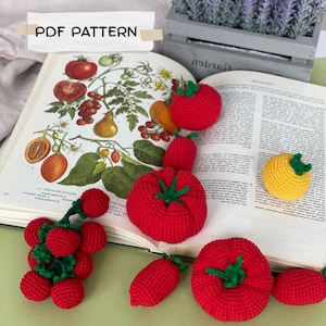 May include: A PDF pattern featuring crochet vegetables including a cluster of red berries, several round red tomatoes, a yellow tomato, and elongated red vegetables, all with green stems, displayed on an open book with botanical illustrations.