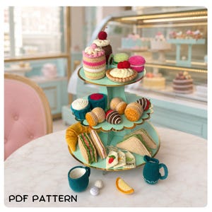 May include: A three-tiered light green display stand holding various crocheted food items including cakes, pastries, sandwiches, and tea accessories. The set features colourful yarn creations like a croissant, tea bags, and a small pitcher. Text reads PDF PATTERN.
