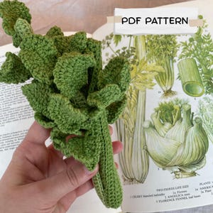 Può includere: Un gambo di sedano lavorato all'uncinetto in verde con foglie testurizzate e un gambo, presentato come modello PDF. L'oggetto è tenuto contro una pagina di libro botanico aperta con illustrazioni di sedano e finocchio con il testo PDF PATTERN.