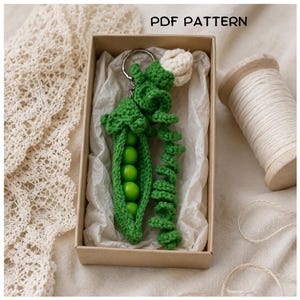 Può includere: Un portachiavi all'uncinetto a forma di baccello di piselli verde con quattro perline sferiche verde brillante all'interno, un viticcio verde arricciato e un piccolo fiore bianco in cima. L'oggetto è in una scatola con il testo PDF PATTERN.