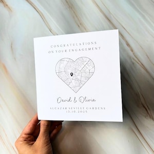 May include: A white engagement card with the text "CONGRATULATIONS ON YOUR ENGAGEMENT". A heart-shaped map design is in the centre, with the names "David & Olivia" below. The card also includes the location and date.