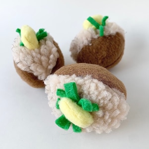 May include: Three brown and white plush stuffed potatoes with green and yellow felt accents. The potatoes are shaped like a traditional baked potato with a dollop of butter and chives on top.