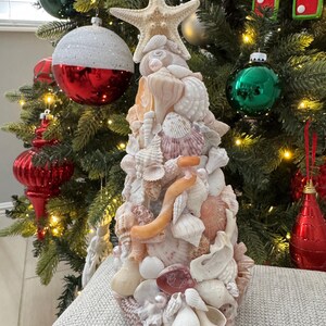 May include: A handcrafted Christmas tree made of various seashells, with a starfish on top. The tree features a mix of white, pink, and beige shells, creating a coastal holiday theme. The tree is approximately 12 inches tall.