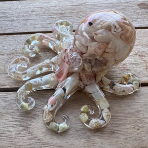 May include: A clear resin octopus figurine filled with various small seashells in shades of white, beige, and pink. The octopus has a rounded head and eight curved tentacles, all containing embedded natural shells visible through the transparent material.