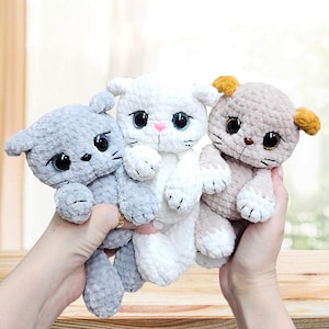 Puede incluir: Tres juguetes de peluche de gato hechos a crochet. El gato de la izquierda es gris, el del medio es blanco y el de la derecha es beige con una oreja amarilla.