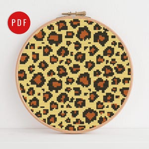 Puede incluir: Un patrón de punto de cruz con un diseño de estampado de leopardo. El patrón está en un aro circular con un fondo marrón claro y manchas negras y naranjas.