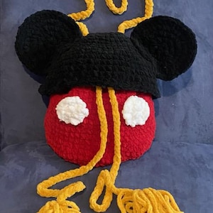 Puede incluir: Una bolsa de ganchillo con forma de Mickey Mouse con orejas negras, un cuerpo rojo con círculos blancos y un cordón largo amarillo con borlas.