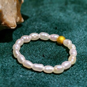 May include: A delicate ring composed of lustrous, off-white oval pearls. A single, small, gold-colored bead accents the design. The ring is set against a dark green, textured background, creating a contrast that highlights the ring's elegance.