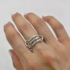 May include: A silver ring with gold accents. The ring features a unique design with interlocking bands. The gold accents are visible on the ends of the ring. The ring is worn on a finger.