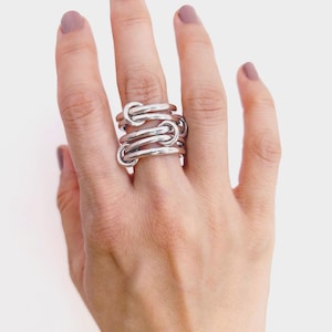 Könnte beinhalten: Ein Stapel silberner Ringe mit einem Design aus verbundenen Knoten. Die Ringe werden an einer Hand mit lackierten Nägeln getragen. Die Ringe haben eine glänzende, metallische Oberfläche und sind zu einem auffälligen Schmuckstück gestapelt.