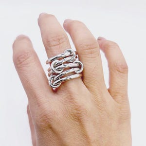 Puede incluir: Un anillo plateado con un diseño único. El anillo presenta múltiples bucles interconectados, creando un aspecto audaz y moderno. El anillo se lleva en un dedo, mostrando sus intrincados detalles y su acabado pulido.