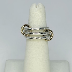 Puede incluir: Un anillo plateado con tres bandas, dos de las cuales están conectadas por detalles circulares dorados. El anillo se muestra sobre un dedo blanco sobre un fondo blanco. El diseño del anillo es moderno y geométrico.