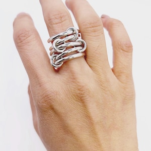 Puede incluir: Un anillo plateado con un diseño de nudo entrelazado único. El anillo está hecho de un metal brillante y presenta múltiples bucles y curvas, creando un aspecto audaz y moderno. El anillo se lleva en un dedo, mostrando sus intrincados detalles.