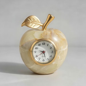 May include: An apple-shaped clock with a gold-colored stem and leaf. The clock face is white with black numerals and gold trim. The apple has a marbled, light green and beige appearance.