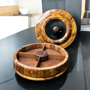 Op de afbeelding: Een tweedelige houten container met een deksel. Het deksel is een groot, rond, gelakt stuk met een donker midden en een kleine, ronde knop. De basis is verdeeld in vier secties. De container is gemaakt van hout met een decoratief bloemmotief.