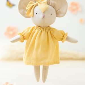 Puede incluir: Una muñeca de ratón suave y afelpada con orejas grandes y redondas y un lazo amarillo. La muñeca lleva un vestido amarillo con mangas con volantes. El ratón tiene una nariz rosa y ojos negros pequeños. Es un juguete para niños.
