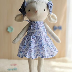 May include: A handmade lamb doll with a cream-colored head and body, wearing a blue floral print dress and matching bows. The doll has a sweet, innocent expression and is standing on a fluffy white surface.
