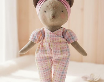 Könnte beinhalten: Handgemachte Bärenpuppe mit rosa Stirnband und kariertem Overall. Die Puppe hat ein hellbraunes Gesicht mit gestickten Details und rosigen Wangen. Der Overall hat ein pastellfarbenes Karomuster. Die Puppe steht auf einer weißen Oberfläche mit Blumendekorationen.