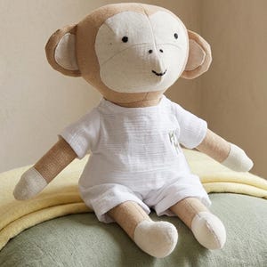 May include: A stuffed monkey toy with tan and cream fabric, featuring embroidered black eyes, nose, and mouth. The monkey wears a white short-sleeved shirt and matching shorts, with a small embroidered detail on the left side of the shirt chest.