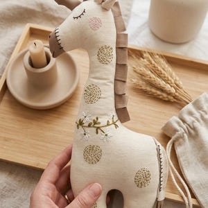 Puede incluir: Un juguete de jirafa de peluche en tela color crema con detalles bordados. La jirafa tiene detalles marrones en las orejas, la melena y las pezuñas. Bordados florales y circulares adornan el cuerpo de la jirafa. Una pequeña bolsa con cordón de color crema está en el fondo.