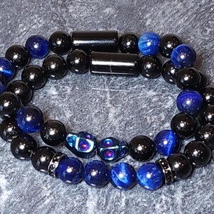Könnte beinhalten: Zwei Armbänder, eines schwarz mit blauen Tigeraugenperlen und das andere mit schwarzen Perlen und zwei Totenkopf-Anhängern.