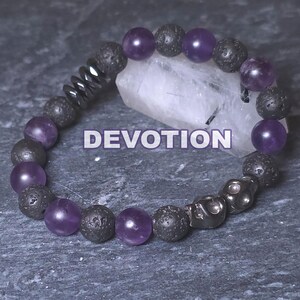 Puede incluir: Una pulsera de cuentas con cuentas de amatista morada y piedra de lava gris oscuro. La pulsera incluye cuentas de calavera gris oscuro y la palabra "DEVOTION" en texto blanco.