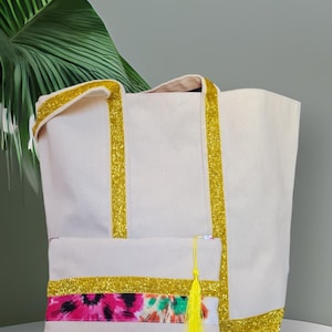 Könnte beinhalten: Ein cremefarbenes Set aus Tragetasche und Clutch. Die Tragetasche hat einen goldglitzernden Besatz an der Oberseite und den Trägern. Die Clutch hat ein farbenfrohes Blumenmuster und eine gelbe Quaste. Das Set ist aus Canvas-Material.