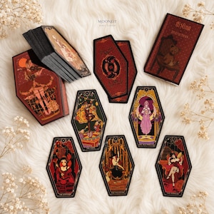 May include: A coffin-shaped tarot deck titled Aisling with a box, instruction booklet, and several cards featuring illustrated figures in red, black, and gold tones. The cards display various characters and symbols, including The Fool and The Magician.