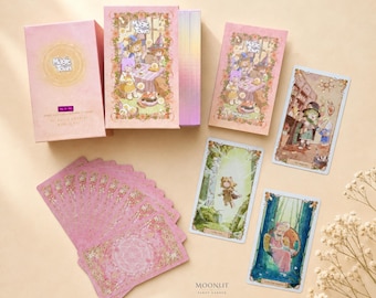 May include: A Magic Town tarot deck set featuring a pink box with gold lettering, a matching guidebook, and several illustrated cards. The cards display whimsical characters in soft pastel colors, with a pink floral pattern on the card backs.