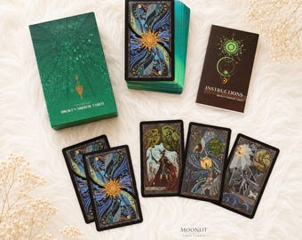 Puede incluir: Un juego de cartas de tarot Broken Mirror que incluye una caja verde, un mazo con diseños de estrellas azules y doradas, un folleto de instrucciones y cuatro cartas individuales con ilustraciones detalladas de figuras y paisajes en tonos oscuros.