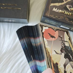 May include: A deck of cards with black edges and a holographic sheen, featuring a card with an illustration of a character, a bat, and a red sun. The deck is positioned next to a dark box with the text "MARIONET" and "AWAKEN IN EMBERS, REBIRTH FROM CHAOS."