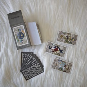 May include: A tarot card deck with a gray box and cards featuring celestial and zodiac-themed illustrations. The box reads "WolsStudios" and "Exploration True Universe". The cards have intricate designs with various colors and symbols.