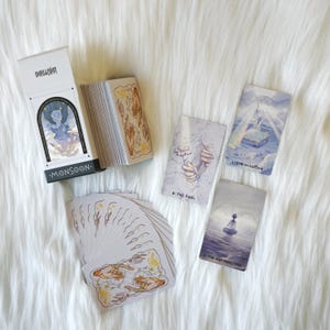 May include: A tarot card deck with a white box featuring the word "Monsoon" and an illustration of a figure. Several cards are fanned out, displaying illustrations of figures and symbols. The cards have a soft, pastel colour palette.