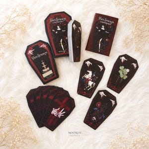 May include: A deck of coffin-shaped Fate Strings Lenormand cards featuring dark, gothic illustrations of figures, a fountain, a horse, and a clover. The set includes a rectangular box and a coffin-shaped box, both decorated with red and black artwork.