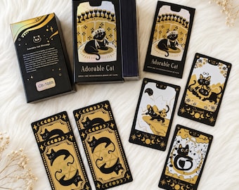 Könnte beinhalten: Ein Kartendeck Adorable Cat Tarot mit schwarz-goldenen Illustrationen von Katzen in verschiedenen Posen. Das Set enthält eine schwarze Schachtel mit dem Text Adorable Cat und CB-1088 sowie einzelne Karten, die The Fool, Strength und The Lovers darstellen.