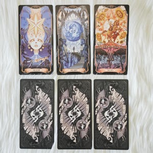 May include: Six rectangular tarot cards arranged in two rows. The top row features The Star, The Moon, and The Sun cards with detailed illustrations. The bottom row displays three identical cards with a dark, intricate design featuring skulls.