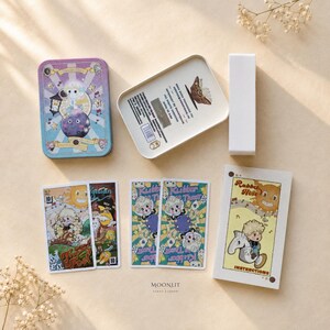 Puede incluir: Un juego de cartas de tarot que incluye una lata rectangular de metal con una ilustración de luna, una tapa abierta con texto impreso, una caja blanca, dos cartas de tarot con arte tipo cómic y un manual titulado Rubber Hose.