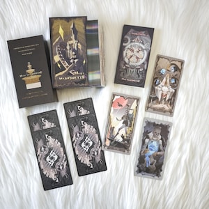 May include: A Null Marionette tarot deck set featuring a black box with a gold fountain illustration, a deck of cards with holographic edges, an instruction booklet, and several individual tarot cards displaying intricate, dark, and surreal artwork.