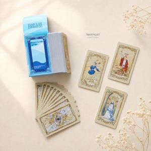Puede incluir: Una baraja de tarot titulada Moonlit Tarot Garden, que incluye una caja azul claro con la etiqueta Resuscitation Potion. El conjunto consta de un mazo de cartas, un abanico de cartas con bordes dorados y tres cartas individuales con ilustraciones detalladas y coloridas.
