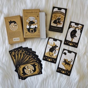 Puede incluir: Una baraja de cartas del tarot "Adorable Cat" con ilustraciones de gatos negros y detalles dorados. El juego incluye una caja, una pila de cartas y varias cartas individuales desplegadas. La caja y las cartas tienen una combinación de colores dorados y negros.