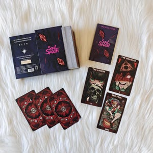 Op de afbeelding: Een Soul Splash tarotdeck met een donkerpaars en roze ontwerp. Het deck bevat een doos, een instructieboekje en verschillende tarotkaarten met unieke illustraties. De kaarten bevatten afbeeldingen van The Lovers, The Magician en The Fool.