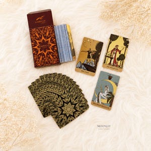 May include: A Sambucus Tarot deck featuring a dark red box with orange mandala patterns, a stack of cards with blue striped edges, and three face-up cards labeled The Fool, The Magician, and The High Priestess with gold and muted color illustrations.
