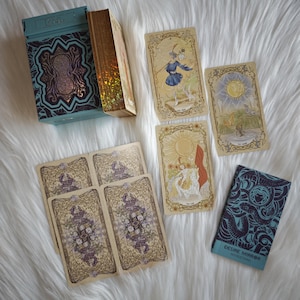 Puede incluir: Una baraja de cartas del tarot con una caja turquesa y cartas con bordes dorados. Las cartas presentan ilustraciones detalladas en tonos beige, dorado y morado. La baraja incluye un folleto con el texto "DESIRE MIRROR INSTRUCTIONS".