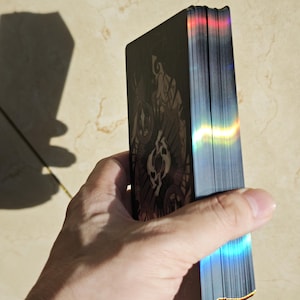 May include: A deck of rectangular playing cards with black backs featuring a dark, intricate pattern. The edges of the cards are finished with a metallic, holographic foil that reflects light in vibrant rainbow colors, creating a shimmering effect.