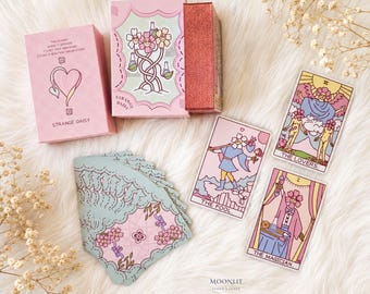 May include: A tarot card deck titled Strange Daisy featuring a pink box with a heart illustration, a box with a floral design, and three cards labeled The Lovers, The Fool, and The Magician. A stack of cards with a light blue and pink floral pattern is included.