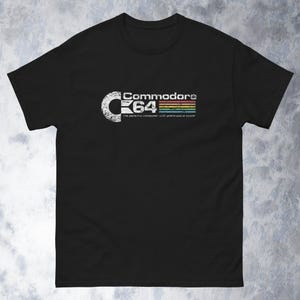Puede incluir: Camiseta negra con el logotipo de Commodore 64 en blanco, con el texto "Commodore 64" y la frase "the personal computer with professional power". El logotipo incluye un diseño de rayas de arcoíris retro.
