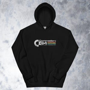 Puede incluir: Sudadera con capucha negra con el logotipo blanco de Commodore 64. El texto "Commodore 64" y el lema "the genuine computer with professional power" se muestran en un estilo retro. El logotipo incluye una franja arcoíris.