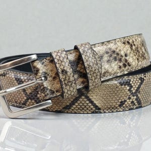 Peut inclure: Une ceinture enroulée avec un motif peau de serpent dans les tons marron, beige et noir. La ceinture a une boucle rectangulaire argentée et un intérieur noir. La ceinture est probablement en cuir.